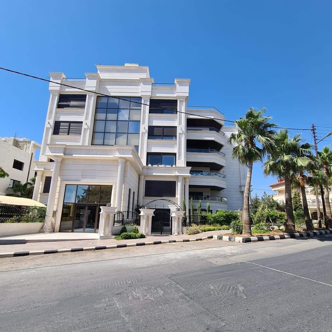 عمارةٌ سكنية بواجهة حجرٍ أبيض وأعمدةٍ ومدخلٍ زجاجيّ، ونخيلٌ أمامها